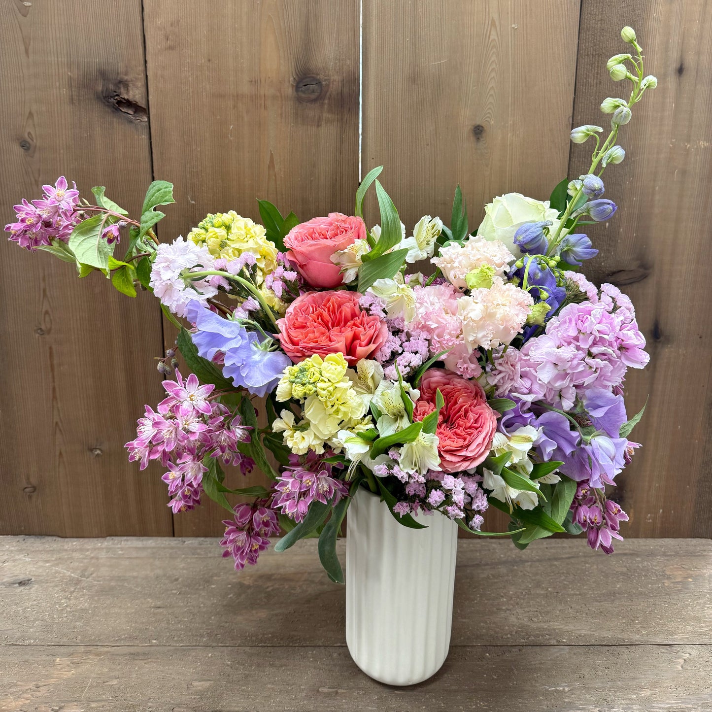 Bouquet of colorful flowers in a white vase on a wooden surface with a wooden fence background.